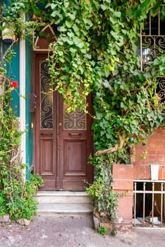 Exterior shot of decorated wooden door beside wrought iron window, mostly covered with green dense climber plants, in a red brick wall, on city street