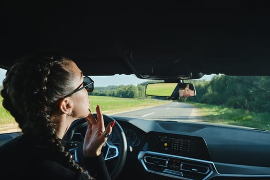 September 12, 2021, Minsk, Belarus - Pretty woman sitting behind the steering wheel, looking to the rearview mirror and fixing lipstick
