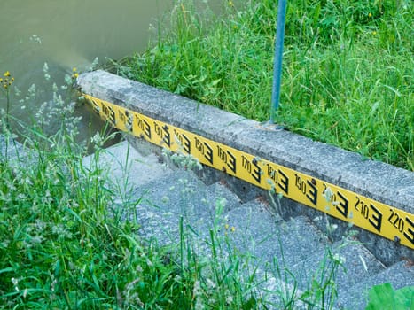 Water level indicator with stone steps and black writing on a yellow background on a grass-lined embankment.