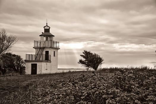 Lighthouse depicted in sepia, Spodsbjerg Fyr in Huntsted on the coast of Denmark. Sun rays shining through the clouds. Meadow with trees. Landscape photo from sea