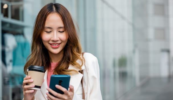 Happy business woman hold mobile phone and paper cup of hot drink outdoor walking on city street, Asian businesswoman holding coffee cup takeaway and smartphone go to work she walking near her office