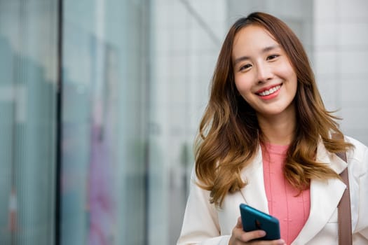 Happy business woman smiling using mobile phone outdoor walking on city street urban looking to camera, Asian businesswoman texting smartphone commuting work she walking near her office building