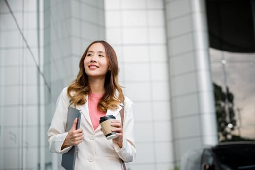 Young business woman wearing white suit jacket and smile while go to work outdoor mirror building background. Confident Businesswoman with cup of coffee holding Laptop walking outside office building