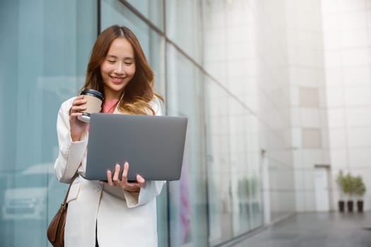 Confident businesswoman using computer laptop standing while drinking a cup of coffee takeaway at the city exterior. Beautiful smile female being ready to work. Business lifestyle concept.