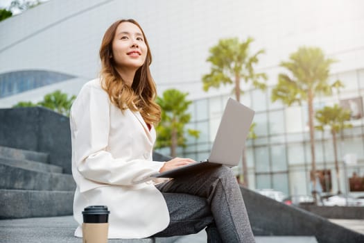 Remote working concept outside the office. Happy young Asian woman freelance smile looking laptop computer with smile while working with coffee cup while sitting on stairs outdoors in the city.