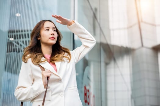 Portrait of confidence young businesswoman standing outside office building in city raise your hand to shade the sun. Happy woman wearing white suit jacket with brown bag at sunlight outdoors.