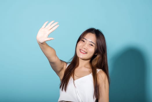 Female raising arm and hand in protection and block sunshine in studio shot isolated on blue background, Asian young woman smile hand up cover face protect sun light and UV, sunscreen cream advertise