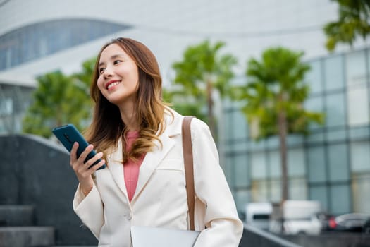 Portrait of confidence Asian businesswoman holding laptop and check message on mobile smart phone. She successfully work go to against city scene of modern office buildings in the city. cellphone