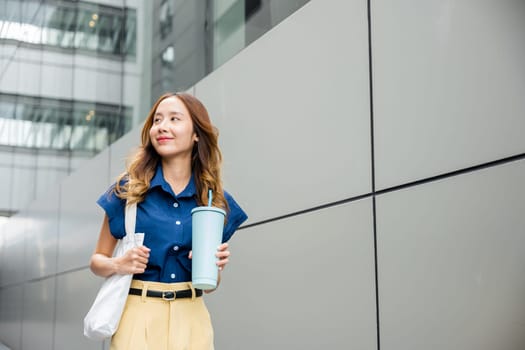 Asian beautiful business woman confident smiling with cloth bag holding steel thermos tumbler mug water glass she walking outdoors on street near modern building office, Happy female