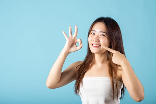 Dental health concept. Asian young woman pointing to her strong white teeth and showing OK sign for good studio shot isolated on blue background, Beautiful female point fingers to smiling mouth