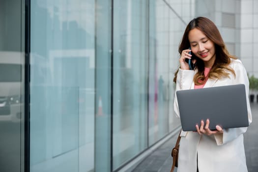 Asian businesswoman working on laptop and talking on cell phone at front building near office, Portrait beautiful business woman in white suit smile discussing issues on smart mobile phone in city