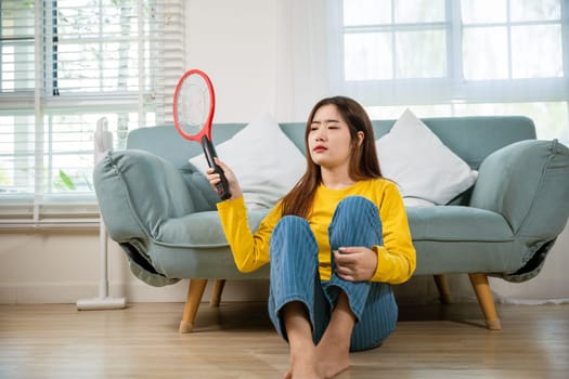 Woman killing mosquitoes hand holding fly swatter like weapon in living room at home, Asian young female sitting floor using mosquito swatter or electric net racket, technology insect killer