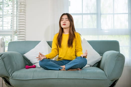 Asian young woman sits on sofa at home and chilling with couch lotus pose eyes closed, female practise yoga and meditation in lotus position, mindfulness lifestyle, happy relaxation leisure people