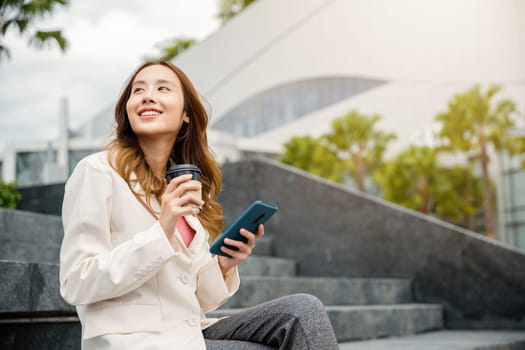 Lifestyle woman business people smiling using mobile smart phones in city office building. Young Asian female wearing blue shirt while use cellphone with holding coffee cup sitting on stair outdoors