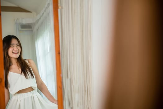 Asian beautiful young woman trying on blouse and smiling while standing in front of mirror in room, Happy female she wear on front mirror looking dress at home after morning wake up