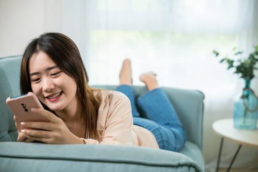 Smiling woman relaxed social online communication on smartphone, Happy Asian female using smart mobile phone lying on sofa in living room room at house, technology gadget online application