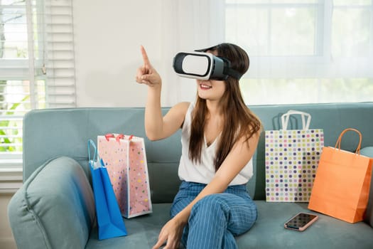 Happy young woman customer wearing 3d VR glasses virtual reality headset with shopping bags around she touching and pointing on air to online shopping in living room at home, digital cyberspace