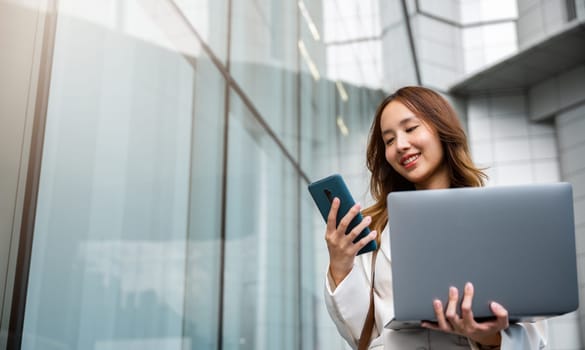 Confident businesswoman using smart phone with computer laptop standing in the city. Beautiful businesswoman being ready to work. Business concept.