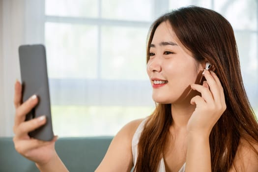 Lifestyle Asian young woman wearing wireless earphones listening to favorite song on her smart mobile phone, Happy relaxed female wear earbuds listen song on smartphone in bright living room at home