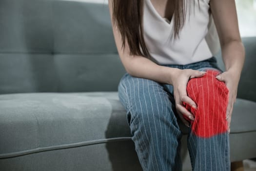 Asian young woman sitting on sofa and feeling suffering knee pain in living room at home, female massage her knee leg pain with painful face, Health bone problems self physiotherapy