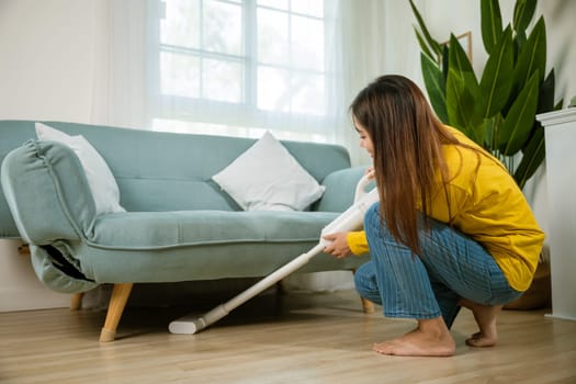 Housewife female dust cleaning floor under sofa or couch furniture with vacuum cleaner, Happy Asian young woman with accumulator vacuum cleaner at home in living room, household and housework concept