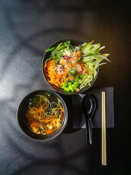 Japanese-style lunch. Bowl with fresh salmon, carrots, cucumbers, green beans, chukka lettuce. Shiro miso soup. Wooden chopsticks and an Asian spoon on a black table at an Asian restaurant.