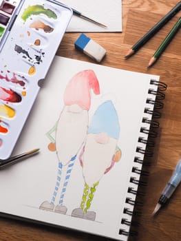 Two gnomes as watercolor drawing to use as Christmas card, hand made seasonal gift card, watercolor artist tools on a wooden table