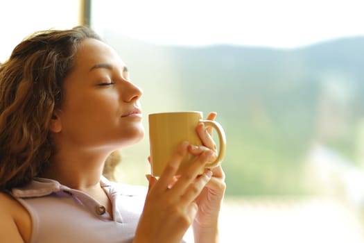 Happy woman smelling coffee relaxing beside a window at home with closed eyes