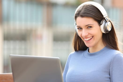Happy woman with headset and laptop looks at you in the street