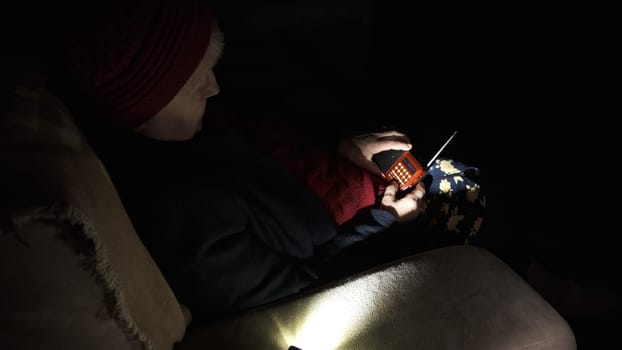 Ukraine: Grandmother holds radio in hands sitting in darkness blackout during russian terroristic attacks. Senior woman in winter clothes in darkness on sofa with hand radio receiver listening news.