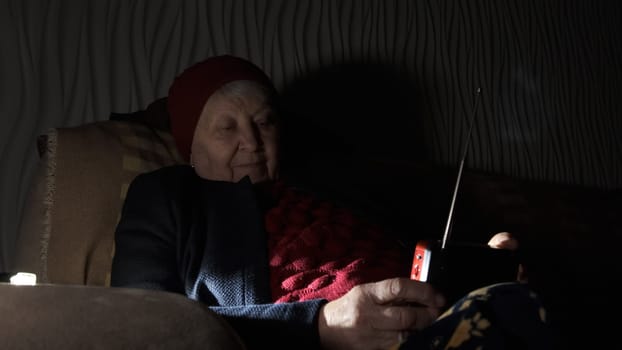 Ukraine: Grandmother holds radio in hands sitting in darkness blackout during russian terroristic attacks. Senior woman in winter clothes in darkness on sofa with hand radio receiver listening news.