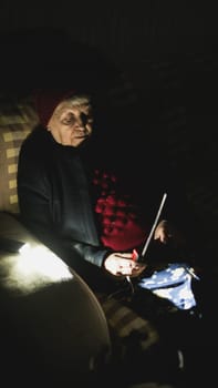 Ukraine: Grandmother holds radio in hands sitting in darkness blackout during russian terroristic attacks. Senior woman in winter clothes in darkness on sofa with hand radio receiver listening news.