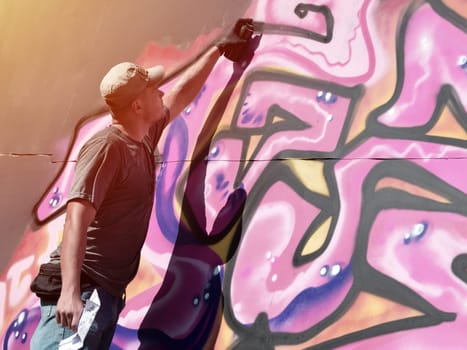 Young caucasian male graffiti artist drawing big street art painting in blue and pink tones. Stylish man in denim shorts and grey t shirt made new graffitti artwork outdoors in bright sunny day