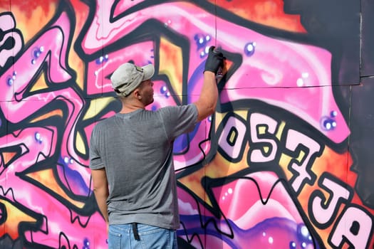 Young caucasian male graffiti artist drawing big street art painting in blue and pink tones. Stylish man in denim shorts and grey t shirt made new graffitti artwork outdoors in bright sunny day