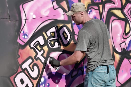 Young caucasian male graffiti artist drawing big street art painting in blue and pink tones. Stylish man in denim shorts and grey t shirt made new graffitti artwork outdoors in bright sunny day