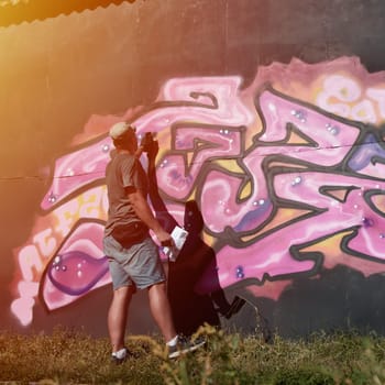 Young caucasian male graffiti artist drawing big street art painting in blue and pink tones. Stylish man in denim shorts and grey t shirt made new graffitti artwork outdoors in bright sunny day