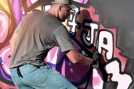 Young caucasian male graffiti artist drawing big street art painting in blue and pink tones. Stylish man in denim shorts and grey t shirt made new graffitti artwork outdoors in bright sunny day