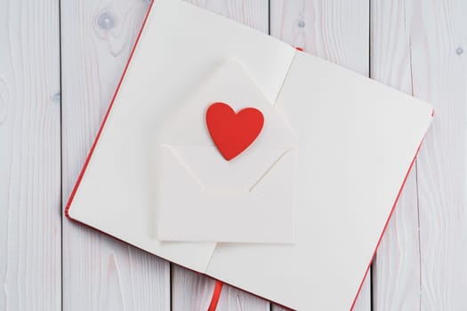 Red diary book with white paper with red heart in craft envelope on light wooden desk background. Sending love letter, message. Falling in love in saint valentines day or 8 march. Copy space, flat lay
