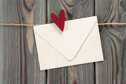 Cute handmade white paper envelope with red heart as stamp, hanging on rope on dark wooden background. Secret present and send feelings on saint Valentine Day. Love letter. Copy space, mockup