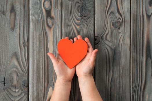 Unrecognizable open hands holding and showing small red paper heart on dark wooden background. I give you my heart and soul. Family and children day or valentines day celebration. Copy space, top view