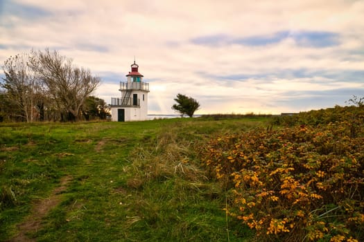 Lighthouse, Spodsbjerg Fyr in Huntsted on the coast of Denmark. Sun rays shining through the clouds. Meadow with trees. Landscape photo from sea