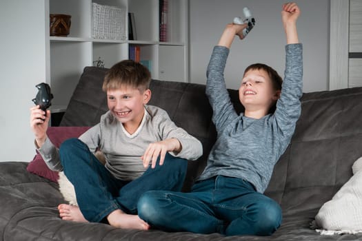 Overjoyed teenagers sit on sofa with game joysticks. Cheerful boys play videogames. Kids having fun together in home room. Modern electronic toys, winning game. Best friends lifestyle.