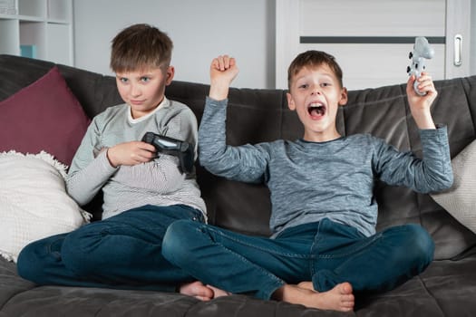 Different emotions from winning and losing. School aged boys sit on sofa with joysticks. Children play videogames. Kids having fun together at home. Game competition. Best friends lifestyle.