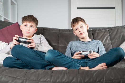 Cute school aged boys sit on sofa with game joysticks. Children play videogames. Kids relaxing in home room at daytime together. Interesting amusement, hobby, electronic toys. Best friends lifestyle.