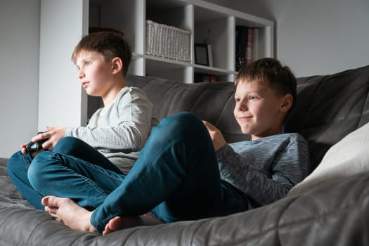 Cute school aged boys sit on sofa with game joysticks, from below view. Children play videogames. Happy kids relaxing in home room at daytime together. Modern electronic games. Best friends lifestyle.