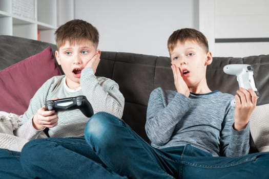 Shocked school aged boys sit on sofa with game joysticks. Children play videogames. Kids relax in home room at daytime together. Disappointment of losing game. Best friends lifestyle.