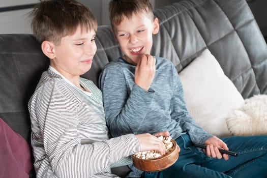 Laughing school aged boys sit on sofa, eat popcorn and watch comic TV show. Overjoyed children relaxing at home. Kids having fun in domestic room at daytime together. Best friends lifestyle.
