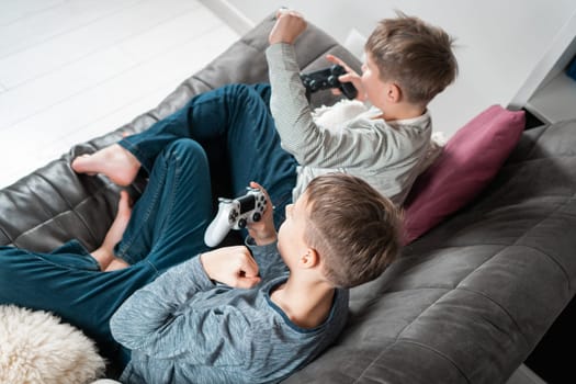 School aged boys sitting on sofa with game joysticks, side view from above. Children play videogames. Kids having fun and relaxing at home. Interesting amusement. Best friends lifestyle.