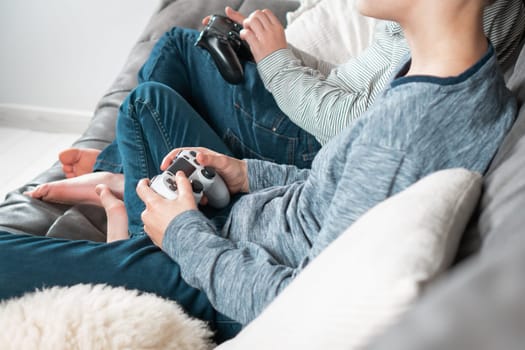 Hands of guys sitting on sofa with game joysticks, side view. Children play videogames. Boys in denim and casual longsleeves relaxing at home. Interesting amusement. Best friends lifestyle.