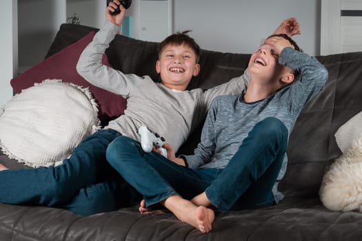 Rejoicing teenagers sit on sofa with game joysticks. Laughing boys play videogames. Kids having fun together in home room. Interesting amusement, winning game. Best friends lifestyle.